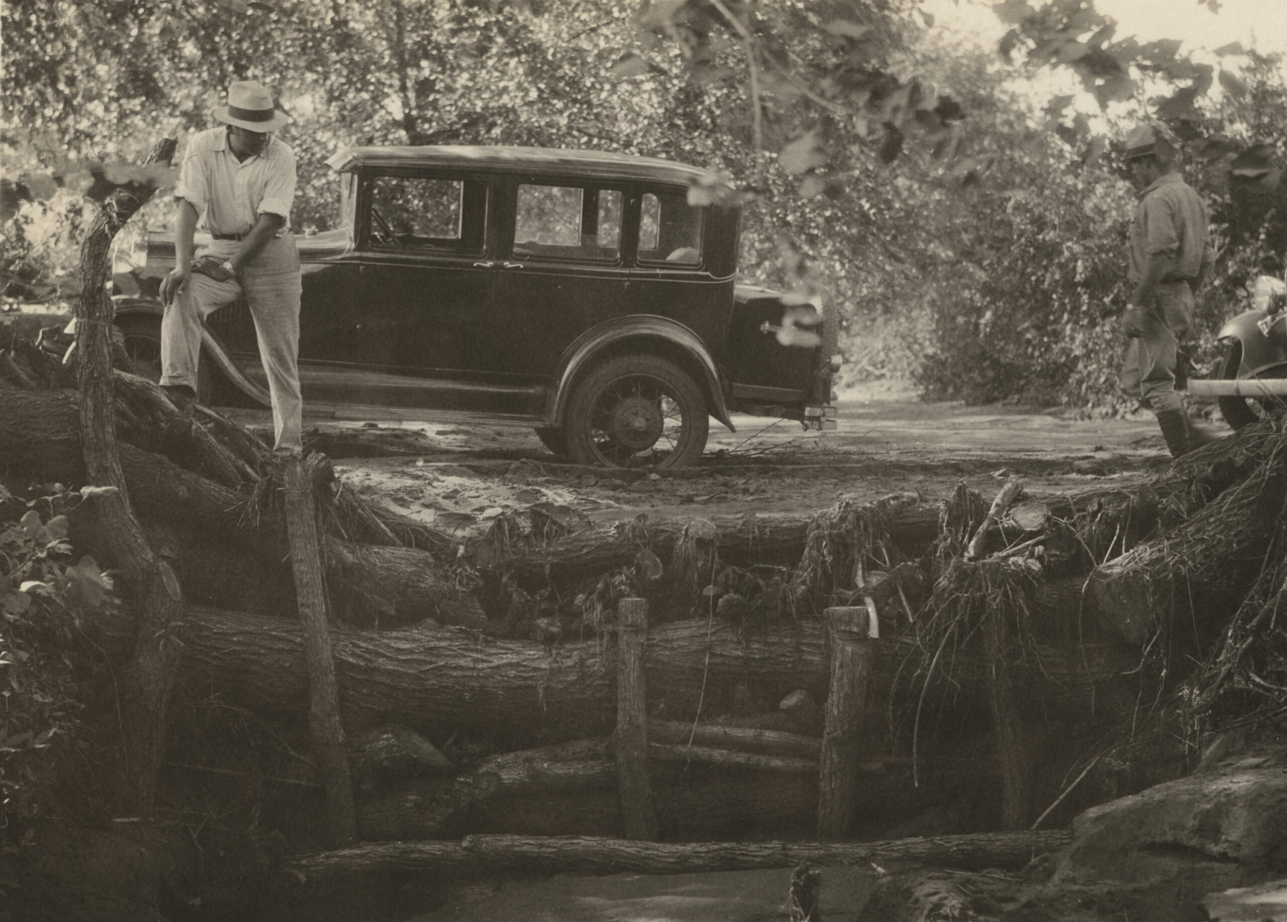 1927 thru 1933 - 2 men with car looking at small log bridge. Binger area, log dam on road.