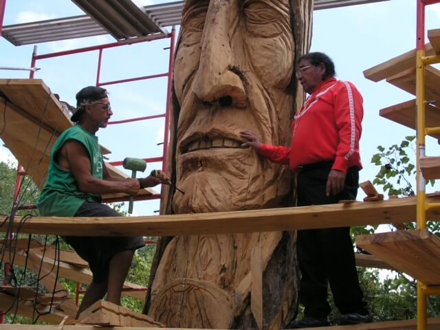 Two people working on wood totem sculpture
